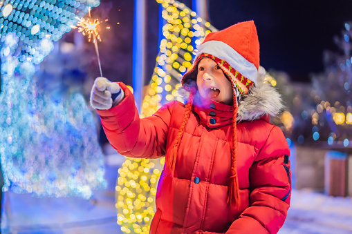 Little boy with sparklers near giant fir tree and Christmas illumination on Christmas market. Xmas holidays on fair.