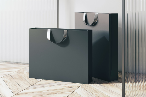 Perspective view on blank black paper shopping bags with place for your brand name or text on light wooden parquet floor near partition and white wall background, close up. 3D rendering, mockup