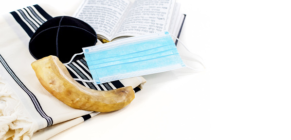 A Covid mask on a prayer shawl with an open prayer book and a shofar horn beside it - Rosh Hashanah concept