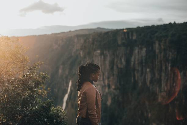 istock Young black woman standing on a cliff next to a waterfall with forest trees at sunset 1464401331