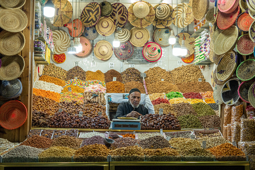 A male vendor stands in his market stall in the Medina (old, historic shopping center) in Marrakech, Morocco. It is evening: electric lights illuminate the stall.
