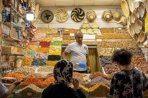 A male vendor stands in his market stall in the Medina (old, historic shopping center) in Marrakech, Morocco. It is evening: electric lights illuminate the stall. In the foreground (seen from the back) two potential buyers