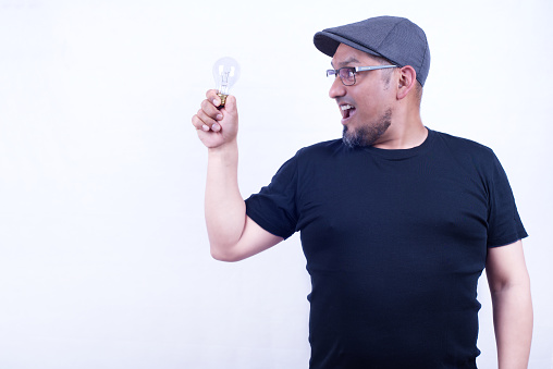 Man on a white background in a cap and glasses, looks at a light bulb in his right hand in a gesture of discovery.