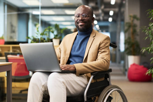 glücklicher junger schwarzer mann mit behinderung sitzt im rollstuhl im büro - accessability stock-fotos und bilder
