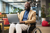 Happy young black man with disability sitting in wheelchair in office