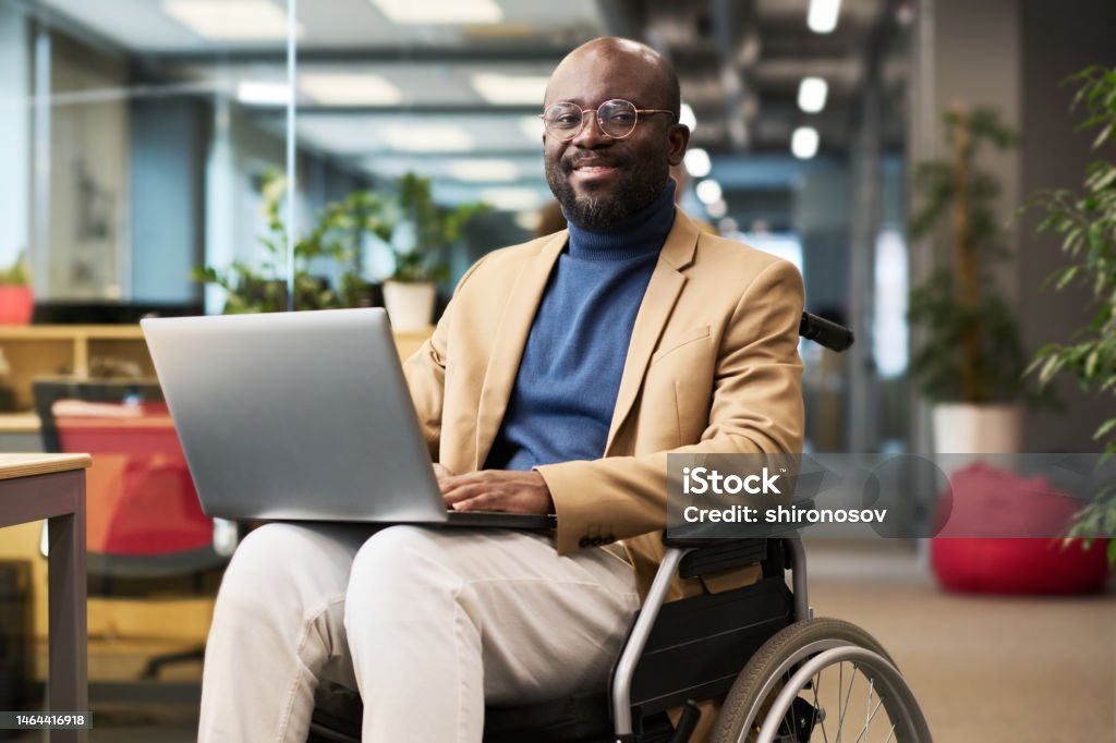 Happy young black man with disability sitting in wheelchair in office Happy young black man with disability sitting in wheelchair in office and looking at camera while analyzing online data on laptop screen Disability Stock Photo
