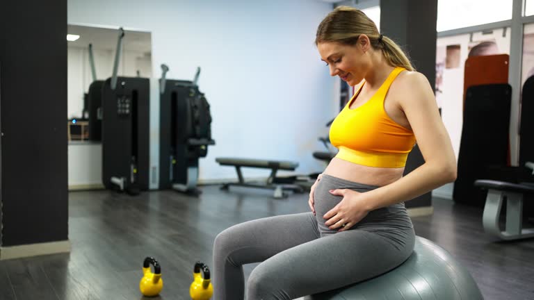 Pregnant Caucasian woman, sitting on fitness ball and touching her pregnant belly