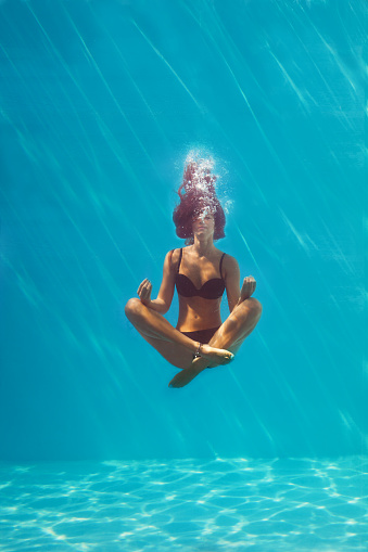 young woman in black bikini in yoga position underwater in diving aquarium, full body shot, front view through the glass