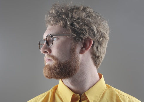 A portrait of an adult ginger man with a beard wearing glasses against a gray background