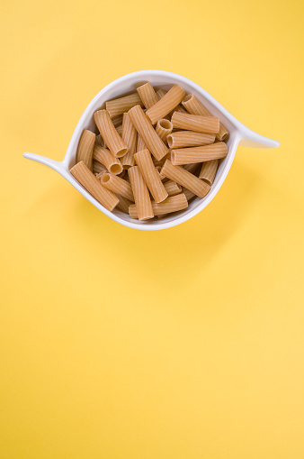 A vertical top view shot of rigatoni in a white bowl on a yellow surface with free space for text