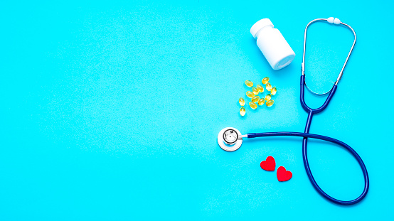 Top view of a blue stethoscope surrounded by some omega 3 capsules, two heart shapes and a pill container. All the objects are at the right side of the image leaving a useful copy space at the left side on a light blue background