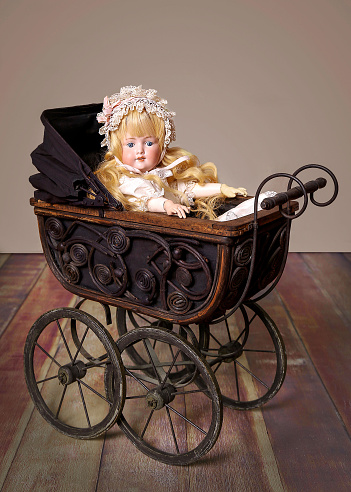 An antique doll with natural white hair sits in a late 19th century doll carriage, dressed appropriately for her age.