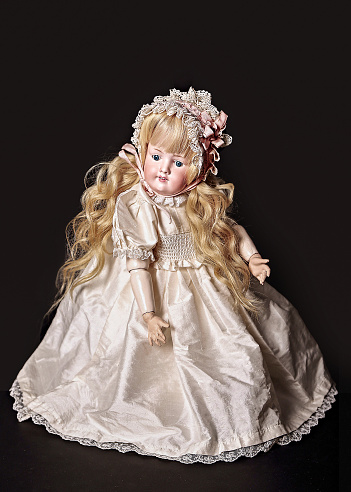 An antique doll with white natural hair sits on an old table in clothes appropriate for her age.