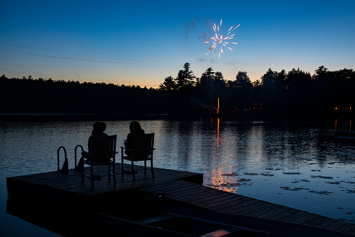 A silhouette of two women sit on a pier at a lake and watch fireworks explode in the night sky on the Fourth of July holiday.