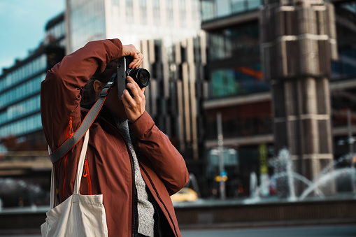 Experienced young photographer takes souvenir photos at sunset in the city. Tourist in red jacket and shoulder bag focuses on the lens. Street photography