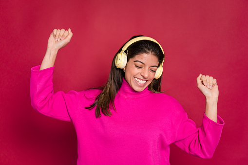 Positive young female in casual clothes smiling and dancing while listening to music in wireless headphones against red background