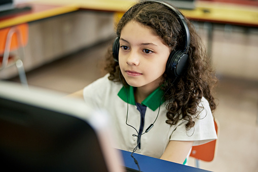 Waist-up view of 9 year old student wearing headphones and looking at desktop pc monitor as she concentrates on her assignment.