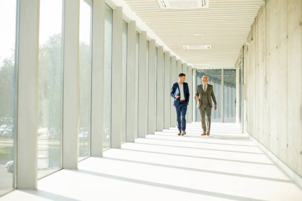 istock Young and a senior businessman walk down an office hallway, deep in conversation 1464444307