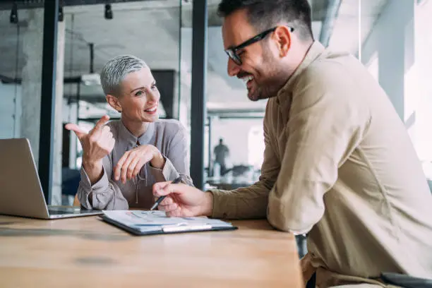 Shot of a two confident business persons sitting on a desk in the office and sharing ideas. Businessman and businesswoman in meeting using laptop and discussing business strategy. Business coworkers working together in the office.