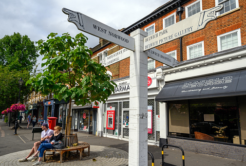 A local sign in Dulwich Village with people and shops. The sign shows directions to Dulwich landmarks and nearby places. Dulwich is in south London, UK