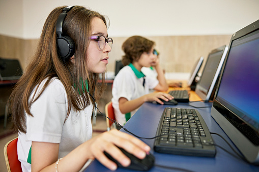 Focus on foreground 11 year old girl with long hair, wearing eyeglasses, headphones, and using mouse as she works on her assignment.