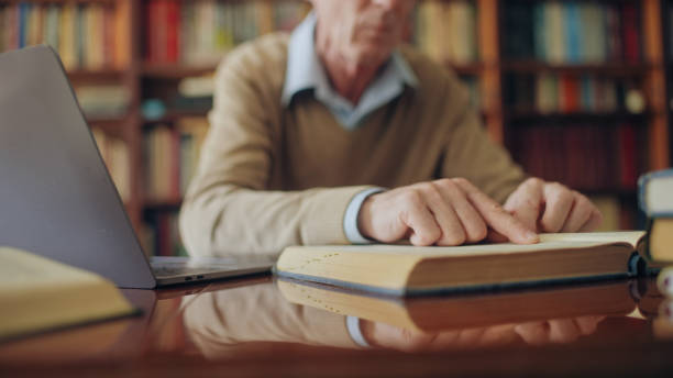 istock Senior historian reading a book and typing notes on laptop, working on an article 1464459447