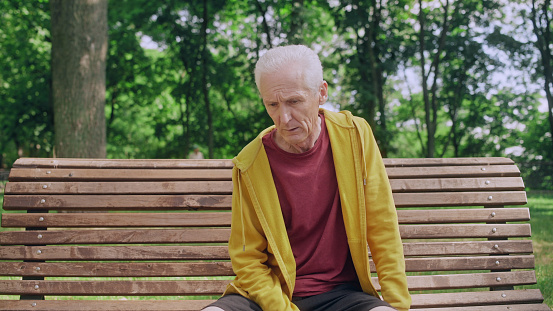 Exhausted senior man sits down on park bench to regain his breath after jogging