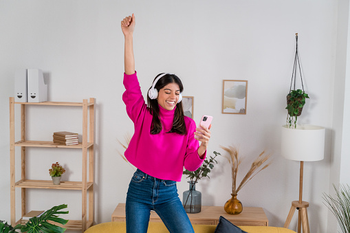 Full body of cheerful young female in casual clothes sitting on yellow sofa and listening to music in wireless headphones