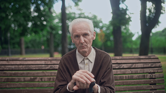 Upset wrinkled senior man sitting in city park alone, holding walking cane