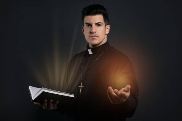 istock Priest in cassock holding Bible with holy light on black background 1464462032
