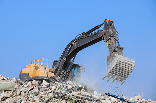 Contemporary excavator with hydraulic press breaks armored concrete leftovers over garbage against cloudy sky at demolition site
