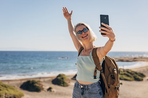 One woman, beautiful female with backpack taking a selfie with smart phone on the beach by the sea.