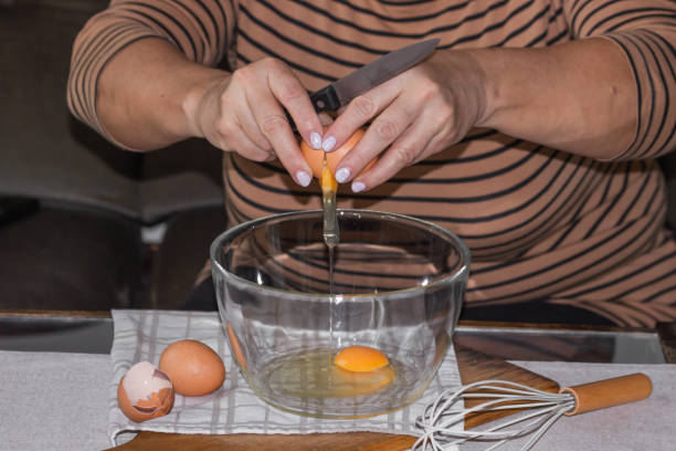 istock Old woman's hands cracking eggs into glass bowl with knife. Prepare for making omelet or pancakes for breakfast. 1464481987