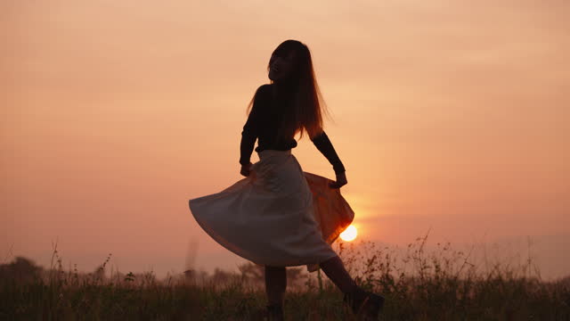 Woman in silhouette holding her skirt and dancing in a meadow in the morning.