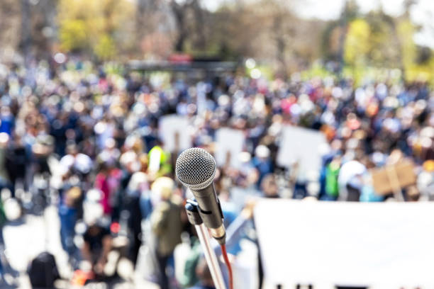 istock Protest, public demonstration, human rights or pension reform strike, focus on microphone, blurred crowd of citizens with flags and banners in the background 1464488460
