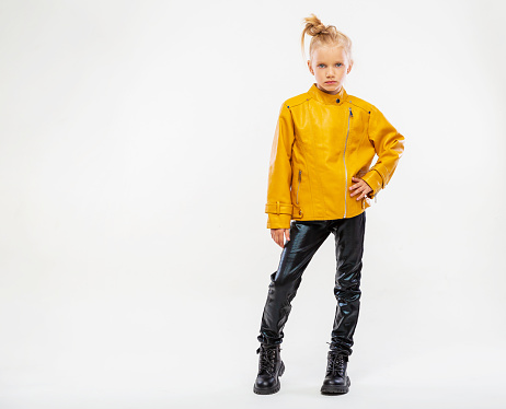 Pretty blonde little girl in a leather mustard jacket, black snake texture leggings and boots. High fashion, full length standing pose, isolated against a studio background