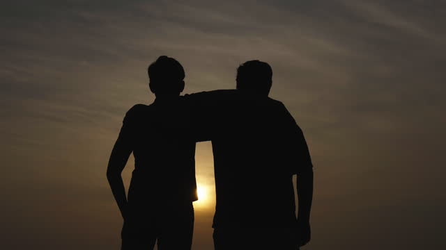 Silhouette of two Asian brothers enjoying view of sunset together as they embrace each other