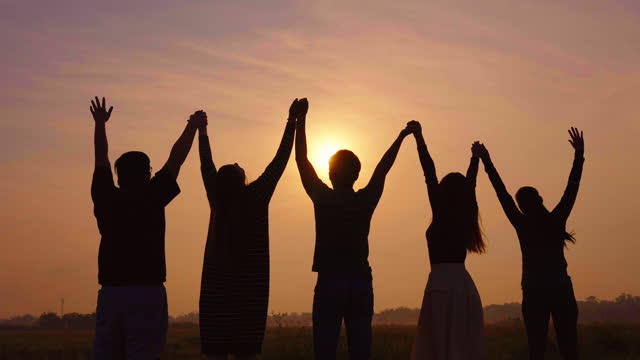 Silhouette of group of five friends raising their holding hands up in the air while the orange sunshine slowly rises