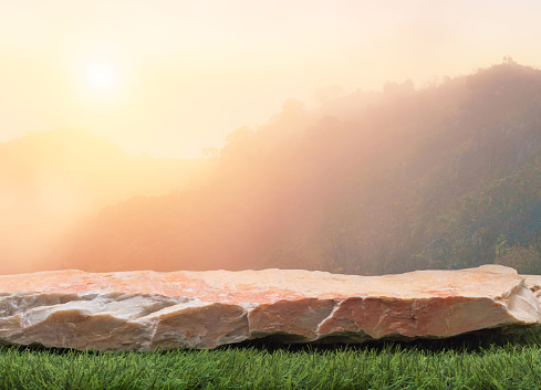 Stone podium table top on fresh green grass with outdoor mountains scene nature landscape at sunrise blur background.Natural beauty cosmetic or healthy product placement presentation pedestal display.