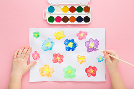 Little child hand holding paint brush and drawing colorful flower heads on white paper on light pink table background. Pastel color. Closeup. Point of view shot. Toddler development. Top down view.