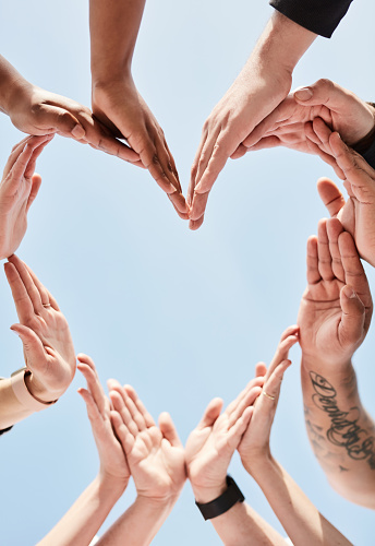 People, hands and unity for heart shape in support, collaboration or teamwork below a blue sky background. Hand of group touching forming love symbol in trust, community or team synergy outside