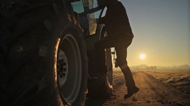 Male farmer climbing into tractor in rural field at sunrise