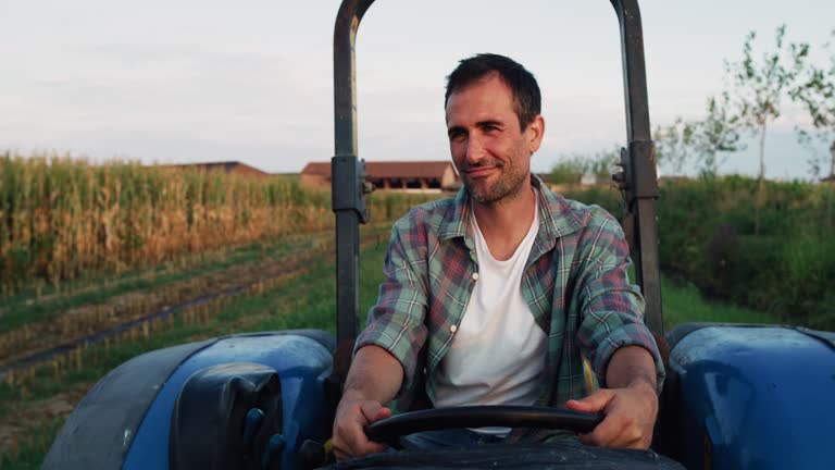 Medium Shot of a Handsome Middle-Aged Man Driving a Tractor on a Road by a Green Field of Corn. Professional Male Farmer Starting his Work Tasks in the Early Morning. He is Checking his Crops