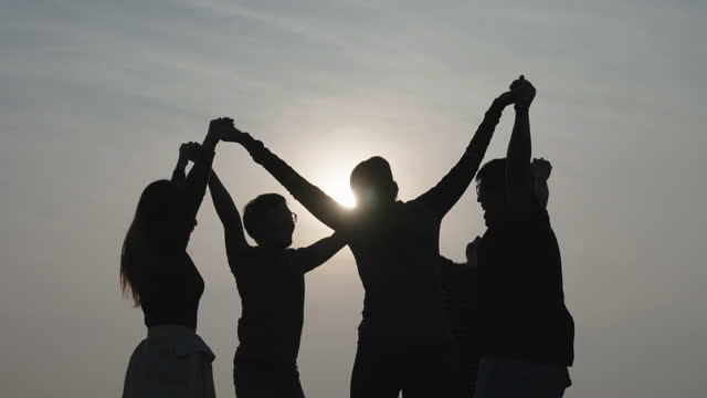 Low angle wide shot of a group of people holding hands and jumping in circle energetically with raising arms at an outdoor team-building camp in silhouette