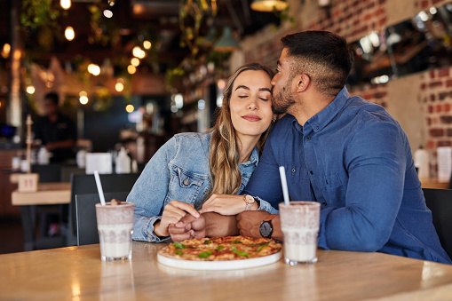 Pizza, kiss and love with couple in restaurant for milkshake, fast food and valentines day date. Celebration, anniversary and sharing with man and woman in cafe for romance, happiness and bonding