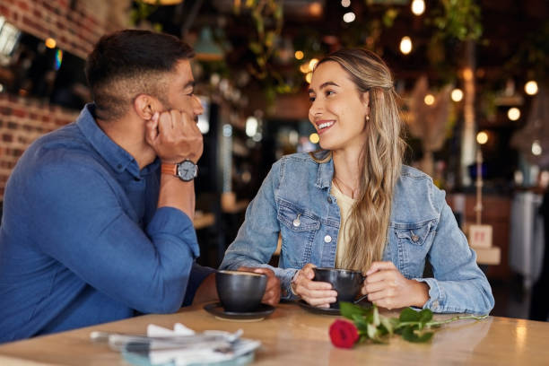 Coffee shop, date and couple of friends with rose for valentines day, anniversary or love in restaurant bokeh. Happy diversity people or woman with love partner at cafe at night for social talking stock photo