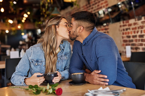 Interracial couple, kiss and coffee shop at breakfast with love, care and happiness. Restaurant, woman and Indian man together in a cafe with morning drink for anniversary kissing in public space