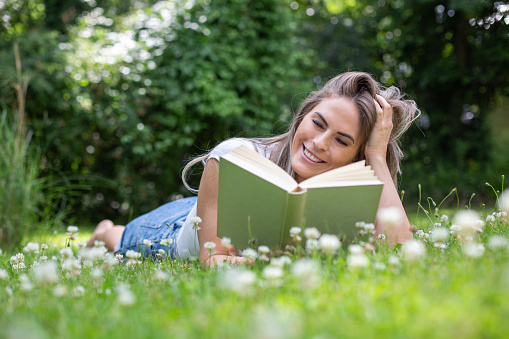 young blond woman lies in the garden between many daisies, reads a book and smiles