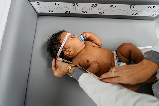 Young female pediatrician examining a newborn baby boy at the pediatric clinic. Measuring his head with measuring tape.