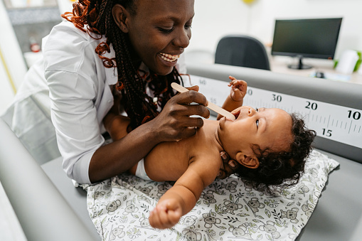 Young black female pediatrician examining a newborn baby boy at the pediatric clinic. Performing a throat exam.